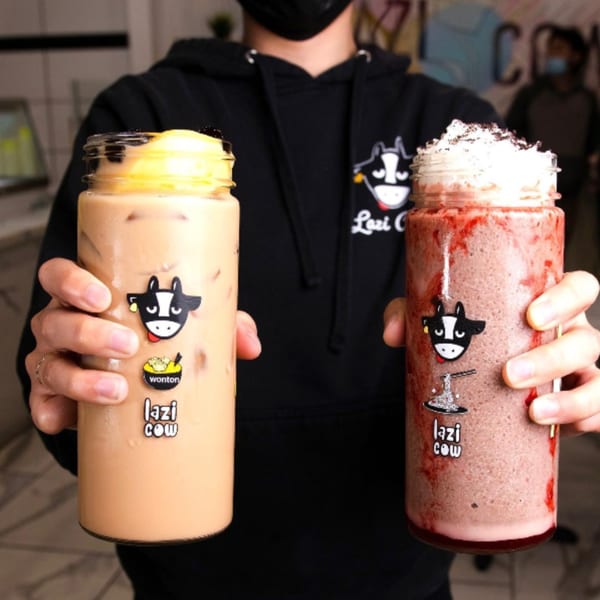 Hands holding two Lazi Cow drinks: a light brown boba tea and a red smoothie with whipped cream.