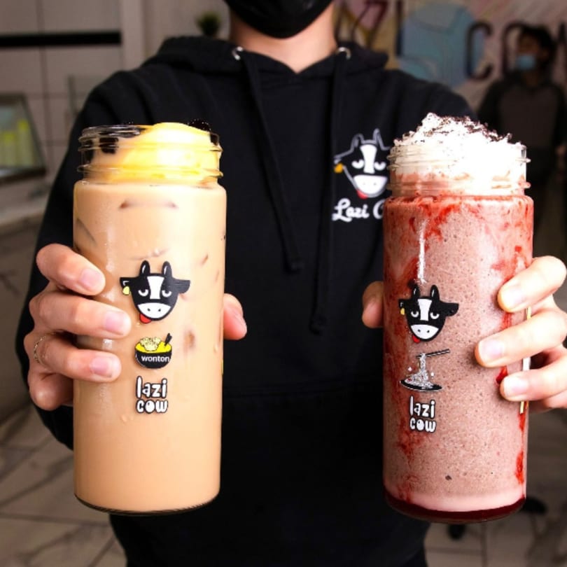 Hands holding two Lazi Cow drinks: a light brown boba tea and a red smoothie with whipped cream.