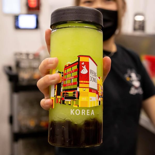 A person holds a Lazi Cow drink with a green top layer and dark bottom layer, featuring a "Korea" label.