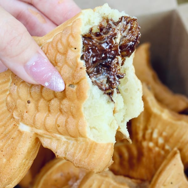 A hand holds a partially eaten taiyaki, revealing a rich chocolate filling inside the fish-shaped pastry.