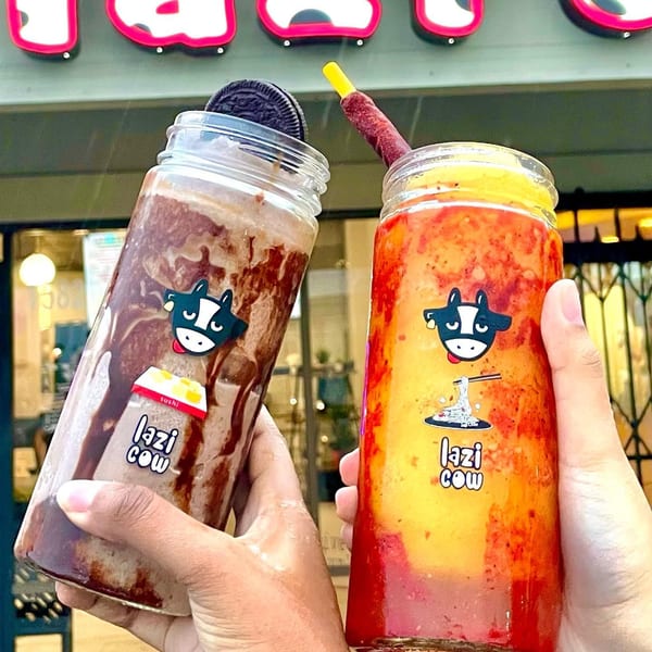 Two hands hold up Lazi Cow drinks, one chocolate with an Oreo, the other a red and yellow fruit drink.
