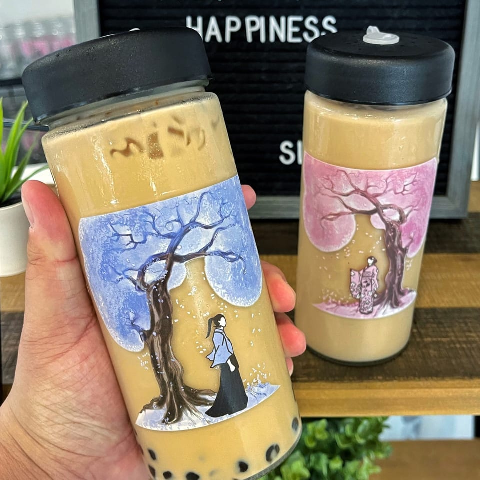 Hand holding a Lazi Cow boba tea with a winter-themed label, another on a shelf.