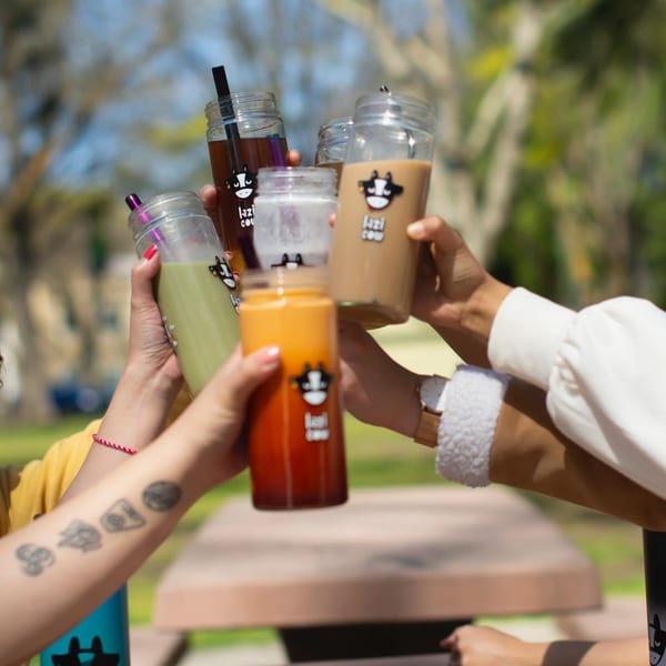 Hands holding up five Lazi Cow drinks in a toast outdoors, with a picnic table in the background.